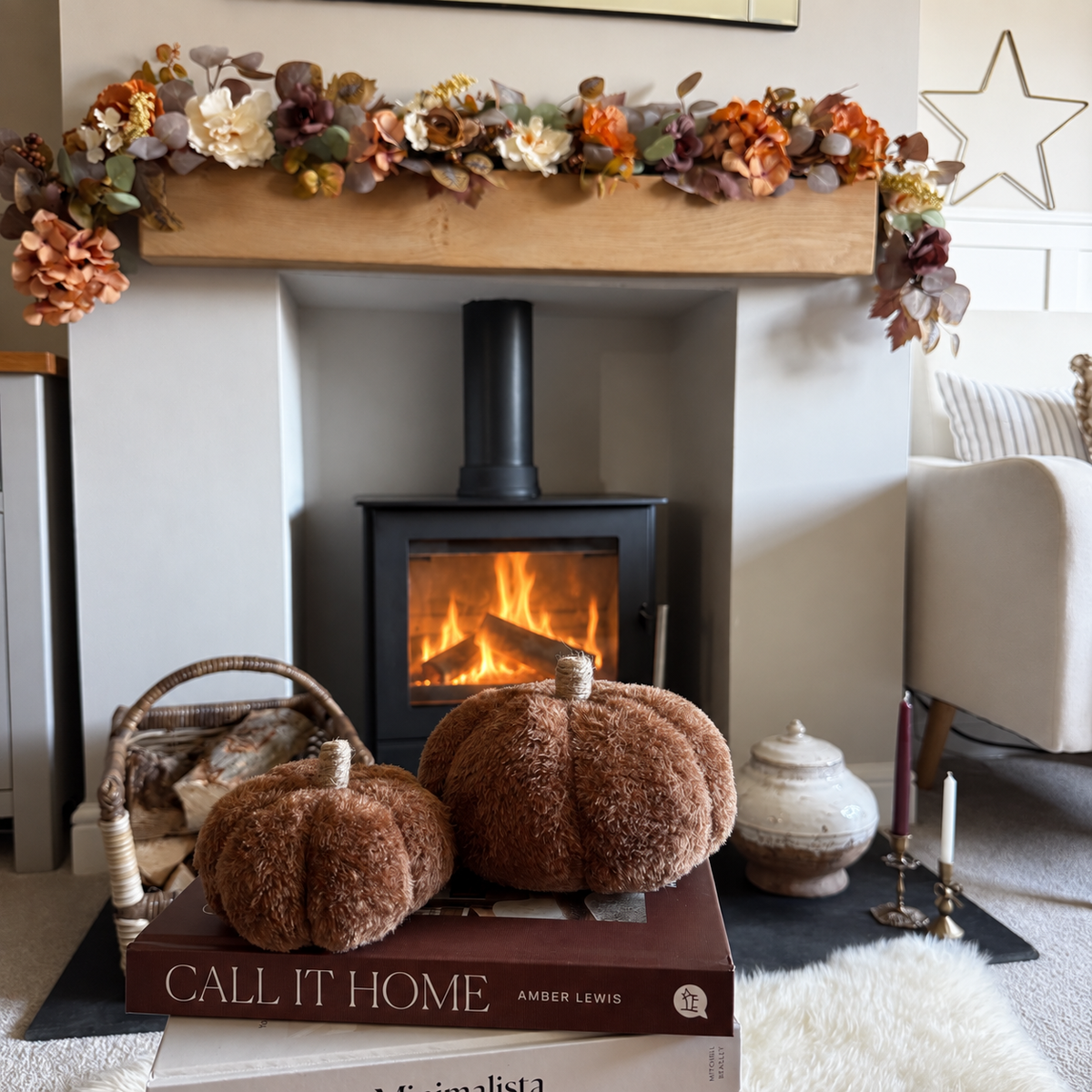 Autumn-themed home decor display featuring two brown fuzzy plush pumpkins and a woven basket of fall flowers on a stack of books in front of a lit wood-burning fireplace.